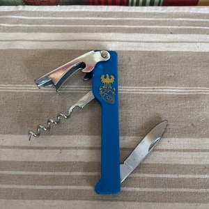 A retro blue plastic and chromed metal pen cutter, bottle opener, corkscrew trio with a gold eagle coat of arms logo