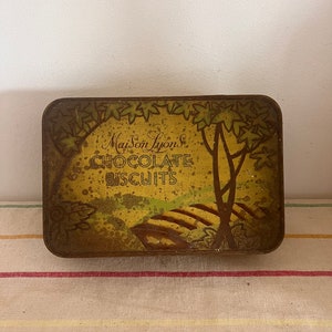 Puede incluir: Una caja de hojalata vintage con fondo amarillo y una ilustración marrón y verde de árboles y un camino. El texto "Maison Lyons Chocolate Biscuits" está impreso en marrón en la caja.