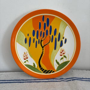 Könnte beinhalten: Dekorative Keramikplatte mit orangefarbenem Rand und farbenfrohem abstraktem Design. Die Platte zeigt einen stilisierten Baum mit blauen Blattformen vor einem gelb-orangen Hintergrund. Zwei kleine rosa Blumen befinden sich am weißen Rand.