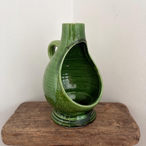 Pode incluir: Um vaso de cerâmica verde com um design aberto único. O vaso tem uma alça e uma superfície texturizada, colocado sobre um banco de madeira. O vaso tem aproximadamente 25 cm de altura.