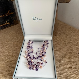 May include: A multi-strand necklace featuring light pink pearls and deep purple faceted beads on thin silver-toned wires, presented in a light grey hinged box with the text Dew Sterling Silver printed on the interior lid.