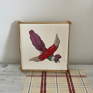 May include: A square ceramic tile with a gold metal frame features a colorful illustration of a bird with red, green, and purple plumage. The bird is flying with its wings spread.