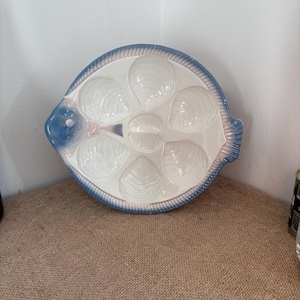 May include: A white ceramic serving platter shaped like a fish, featuring six shell-shaped indentations for oysters. The platter has a blue-tinted rim with a textured, ribbed pattern and a blue fish head detail on the left side of the plate.