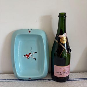 May include: A light blue rectangular dish with a cartoon figure in a red jacket and the text "BORN 1820 - STILL GOING STRONG". Next to it is a green champagne bottle with a pink label.