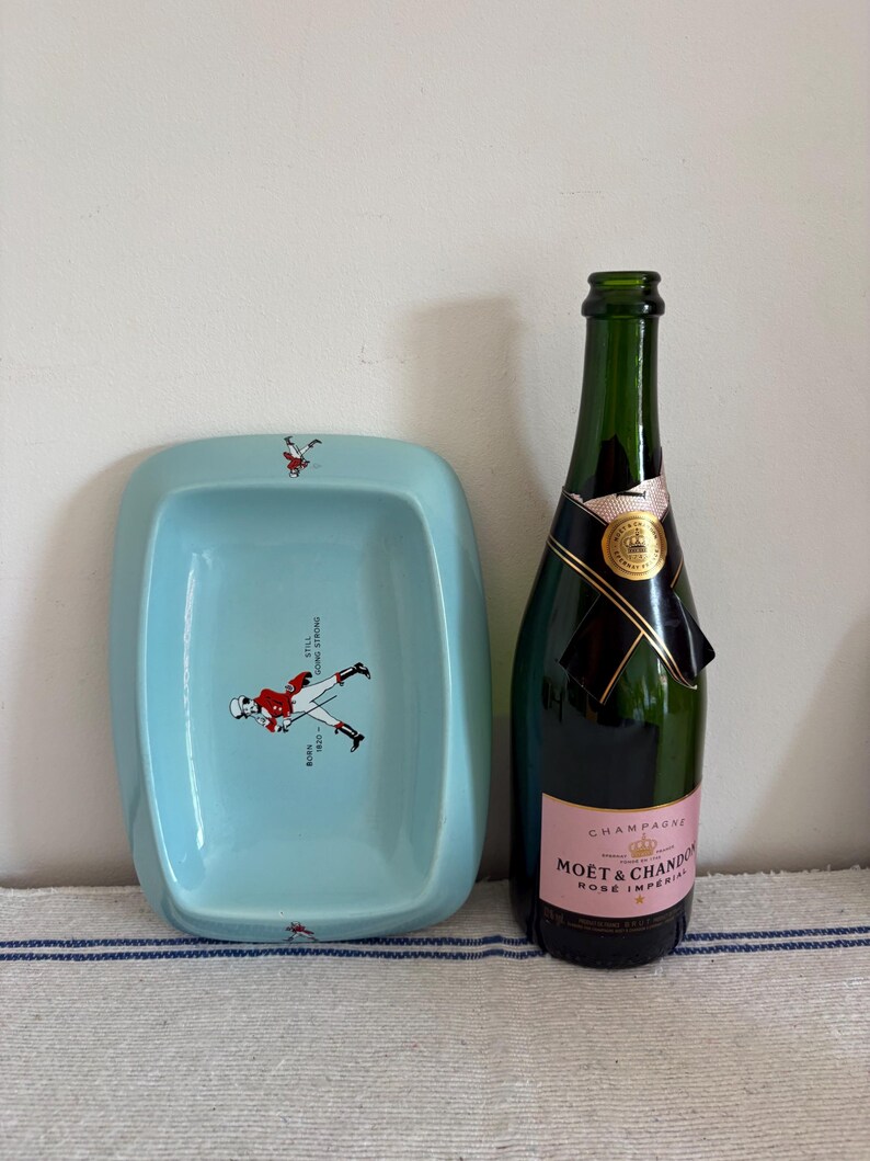 May include: A light blue rectangular dish with a cartoon figure in a red jacket and the text "BORN 1820 - STILL GOING STRONG". Next to it is a green champagne bottle with a pink label.