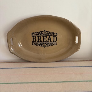 May include: A tan oval bread dish with the word "BREAD" in black, decorative lettering. The dish has two handles and a raised rim. The dish is on striped fabric.