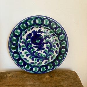 Puede incluir: Un plato redondo de cerámica con un diseño floral. El plato presenta una base blanca con motivos florales azules y verdes. El borde está decorado con un patrón repetitivo de círculos azules y verdes. El plato está sobre una superficie de madera.