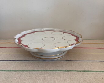 Puede incluir: Soporte para pasteles de cerámica blanca con borde festoneado y borde rojo. El soporte tiene un ribete dorado y una base de pedestal. La superficie superior presenta seis hendiduras circulares. Una pieza decorativa para servir postres.