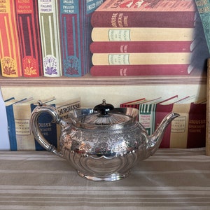Könnte beinhalten: Eine silberne Teekanne mit einem verzierten Design und einem schwarzen Knopf auf dem Deckel. Die Teekanne steht auf einer gestreiften Tischdecke vor einem Bücherregal mit Büchern in verschiedenen Sprachen.