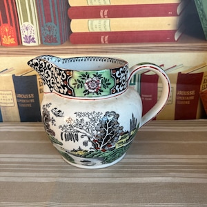 Könnte beinhalten: Eine weiße Keramik-Milchkanne mit einem schwarzen, grünen und roten Blumendesign. Die Kanne hat einen Griff und einen Ausguss.