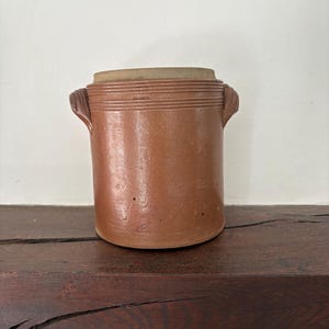 May include: A cylindrical brown stoneware crock featuring a smooth, glazed finish, a slightly recessed top rim, and two small, molded side handles. The vessel has a uniform, earthy tone with subtle surface texture and a sturdy, flat base design.