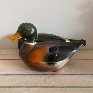 May include: A ceramic duck-shaped container with a green head, orange beak, and brown and black body. A green bottle is behind the duck. The duck is on a striped fabric.