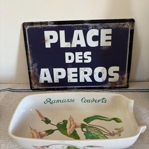 May include: A rectangular white ceramic serving dish with a floral design and the text "Ramasse Couverts" in green. Behind it is a blue sign with white text that reads "PLACE DES APEROS".