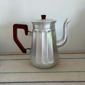 以下が含まれることがあります： 茶色の木製の取っ手と濃い茶色のプラスチック製の蓋が付いた、銀色のアルミ製のコーヒーポット。ポットは溝のあるデザインで、ストライプのテーブルクロスの上に置かれています。