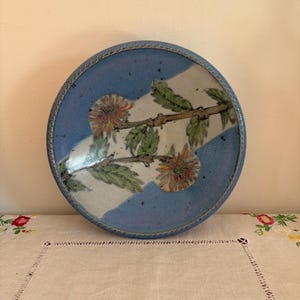 May include: A blue ceramic plate with a white and green floral design. The plate has a rope-like border.
