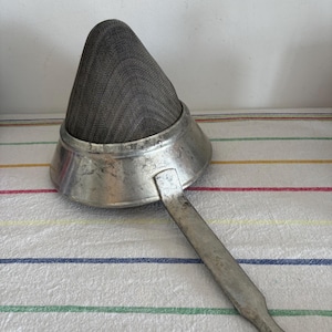 Puede incluir: Un colador de metal plateado con un filtro de malla en forma de cono y un mango largo. El colador muestra signos de desgaste. El mango tiene un agujero en el extremo para colgarlo. Probablemente se utiliza para colar líquidos o sólidos en la cocina. Mide unos 20 cm de largo.