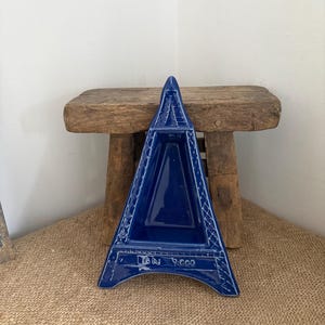 Puede incluir: Un plato de cerámica azul cobalto brillante con forma de torre Eiffel. La estructura presenta un centro triangular hundido y una textura de celosía a lo largo de los bordes. La base del plato tiene grabado el texto "LAN 2000" en letras blancas.