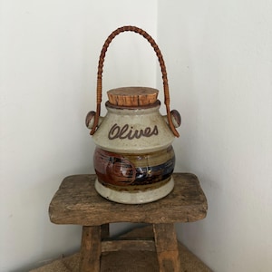 May include: A ceramic jar with a speckled beige finish and the word Olives written in brown script. The jar features a cork lid, a brown woven handle, and a wide band of dark brown and black glaze around the center with a circular accent.