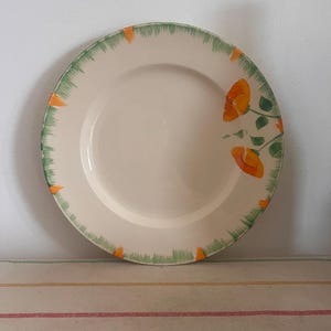 May include: A white ceramic dinner plate with a green and orange floral design. The plate has a scalloped edge and features a stylised poppy flower design.