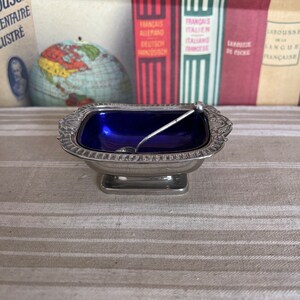 May include: A silver salt cellar with a blue glass liner and a small silver spoon. The salt cellar is sitting on a striped tablecloth.