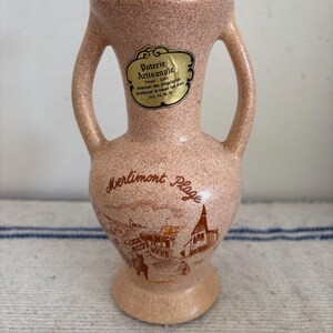 May include: A speckled, peach-colored ceramic vase with two handles. The vase features a gold label with French text and a detailed illustration of a coastal town, including the text "Merlimont Plage".