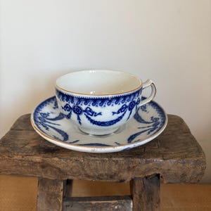 May include: A white teacup and saucer set with a blue floral design and gold trim. The teacup sits on a matching saucer, both resting on a rustic wooden stool. The teacup has a handle and a delicate pattern.