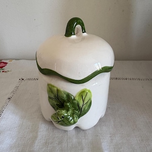 May include: A white ceramic jar with a lid and a green handle. The jar is decorated with green leaves and a small frog. The jar is sitting on a white surface.