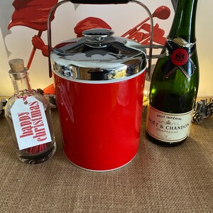 Puede incluir: Una cubitera roja con asa y tapa plateadas, junto a una botella de champán y una pequeña botella de vidrio con una etiqueta de "feliz navidad". Los objetos están sobre una superficie de arpillera.