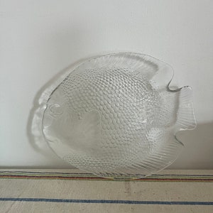 May include: Clear glass serving plate in the shape of a fish. The plate has a textured surface with a scale pattern. The fish plate has fins and a tail, and is suitable for serving appetizers or small dishes.