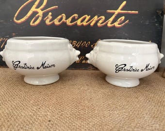 May include: Two white ceramic soup bowls with handles, each inscribed with "Gratinée Maison" in black script. The bowls sit on a burlap surface, with a vintage "Brocante" sign in the background.