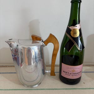 Puede incluir: Una jarra plateada con asa y tapa de madera, junto a una botella de champán verde con una etiqueta rosa que dice "Moët & Chandon Rosé Impérial". La jarra está sobre un mantel a rayas.