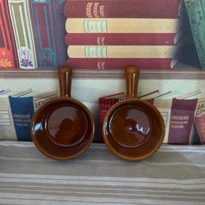 Może przedstawiać: Dwa brązowe ceramiczne ramekiny z zaokrąglonym brzegiem i uchwytem po obu stronach. Ramekiny znajdują się na wzorzystej serwetce.