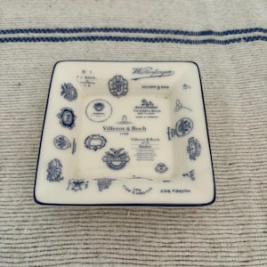 Puede incluir: Plato cuadrado de cerámica blanca con borde azul y diseños azules intrincados. El plato presenta texto y logotipos, incluyendo "Villeroy & Boch 1748". El diseño recuerda a la porcelana vintage.