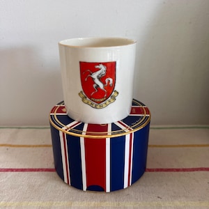 Puede incluir: Una taza de cerámica blanca con un escudo rojo y dorado que presenta un caballo blanco. La taza está sobre una caja azul y roja con un borde dorado.