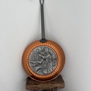 May include: A round copper pan with a long black metal handle. The center features a circular silver-toned relief plaque depicting a blacksmith working at an anvil. The rim of the pan is decorated with a repeating embossed pattern of small circles.
