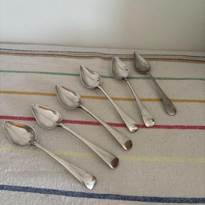 May include: Six silver spoons with oval bowls and long, slender handles are arranged on a striped fabric. The spoons are angled, showcasing their polished surfaces. The fabric has blue, red, yellow, and green stripes.
