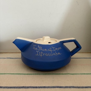Puede incluir: Tetera de cerámica azul con tapa y asa blancas. La tetera tiene las palabras "The Blue of Devon Ilfracombe" escritas en el lateral en letras doradas.