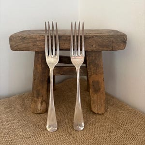 May include: Two silver forks with four tines each, displayed on a small, weathered wooden stool. The forks have a simple, elegant design with a rounded base. The stool is made of aged wood, adding a rustic touch to the scene.
