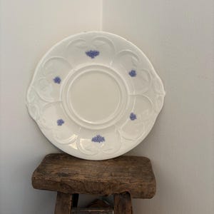 May include: White porcelain plate with a scalloped edge and embossed floral design. The plate features small, light purple floral accents. The plate is resting on a small, rustic wooden stool.