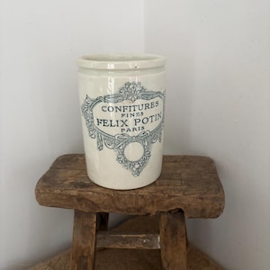 May include: A vintage white ceramic jar with blue text and floral designs. The jar reads "CONFITURES FINES FELIX POTIN PARIS." It sits on a small, rustic wooden stool. The jar is cylindrical with a slightly flared rim.