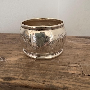 May include: A silver napkin ring with an engraved floral design. The ring is cylindrical with a slightly flared top and base. The metal reflects light, and the ring sits on a wooden surface.