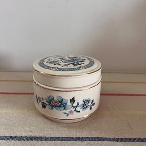 Peut inclure: Un pot en céramique blanche avec un motif floral. Le couvercle est orné d'une bordure dorée et d'un motif floral bleu et blanc. Le pot est orné d'une bordure dorée autour du haut et du bas.