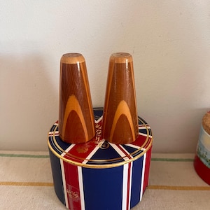 Puede incluir: Un juego de dos saleros y pimenteros de madera en una caja con estampado de Union Jack. Los saleros están hechos de madera clara y oscura con un acabado natural.