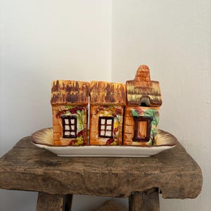 Puede incluir: Un conjunto de tres recipientes de cerámica con forma de casa, con tejados de paja marrones y paredes naranjas, colocados sobre una bandeja ovalada a juego. Cada casa tiene una ventana con una rejilla blanca y está decorada con detalles florales verdes y rojos.