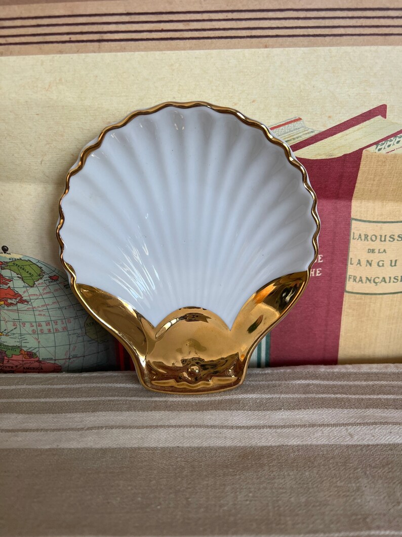 May include: A white porcelain seashell dish with a gold rim and a gold interior. The dish is perfect for holding small items like jewelry or keys.