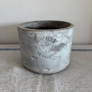 May include: A small, gray concrete planter with a slightly textured surface. The planter is cylindrical with a wide opening and features embossed butterfly designs and the text "FLUTTER BY".