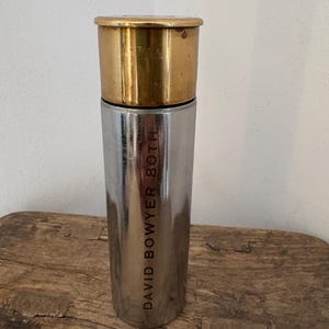 Könnte beinhalten: Ein zylindrischer Metallbehälter mit einem polierten silberfarbenen Körper und einer kontrastierenden goldfarbenen Kappe. Die Seite des silbernen Körpers ist mit dem vertikalen Text DAVID BOWYER 80TH in einer serifenlosen Schrift graviert.