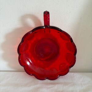 Könnte beinhalten: Eine leuchtend rote Glasschale mit Griff. Die Schale hat einen gewellten Rand und ein kreisförmiges Design in der Mitte. Der Griff ist klein und rechteckig. Die Schale ist wahrscheinlich Vintage oder antik.