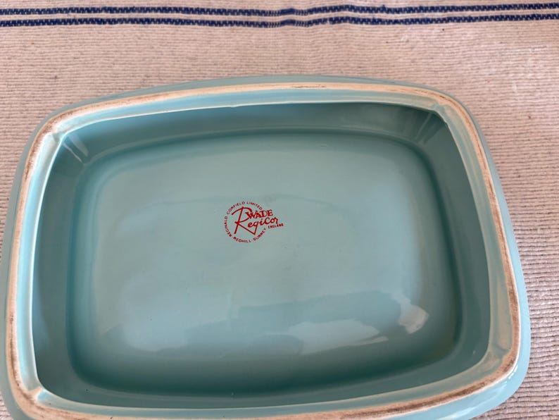 May include: A rectangular, light turquoise ceramic dish with a slightly raised rim. The dish has a glossy finish and a red stamp that reads "MADE RegicO ENGLAND". The dish is likely vintage.