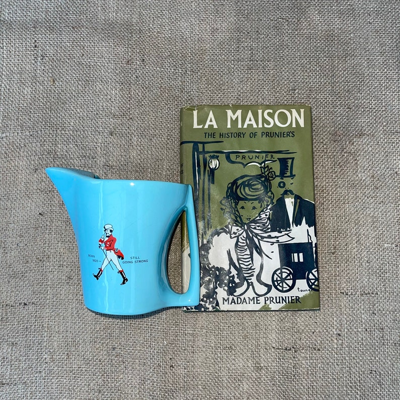 May include: A blue ceramic jug with a red and white illustration of a man walking with a cane and the text "Still Going Strong". The jug is next to a green book titled "La Maison The History of Prunier's" with a black and white illustration of a woman and a man.